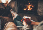 Woman in woollen socks by the fireplace. Unrecognisable relaxes by warm fire with a cup of hot drink and interesting book, warming up her feet. Cozy atmosphere. Winter and Christmas holidays concept.
